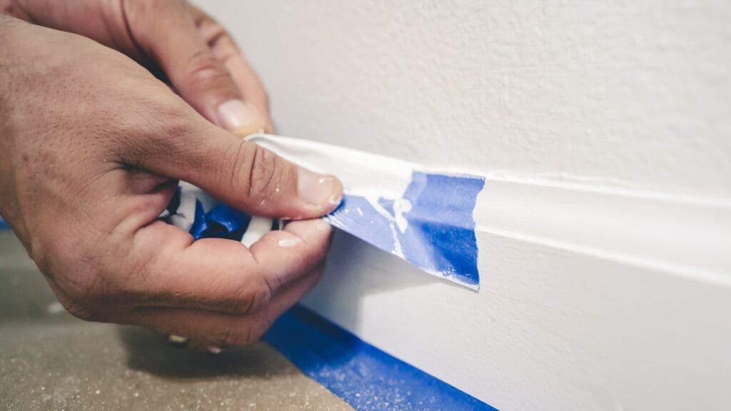 A close up of a person's hand peeling off blue tape after use on a white skirting board.