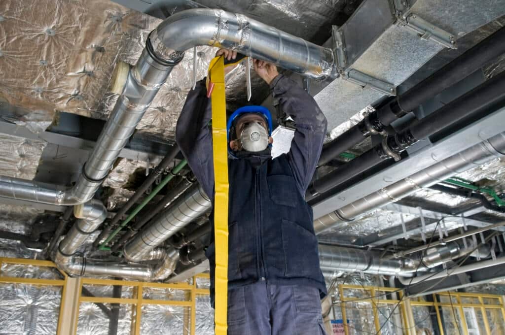 Worker in protective gear inspects industrial pipes on a ceiling, using JTAPE'S "110° High Performance UV Masking Tape". The setting is a metallic, insulated space, conveying focus and precision.