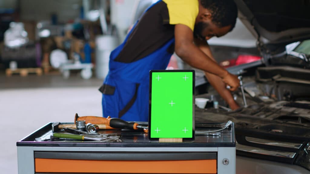 Mechanic in car service uses wrench to replace parts inside vehicle with green screen tablet in front. Expert utilizes professional tools to adjust malfunctioning automobile next to mockup device