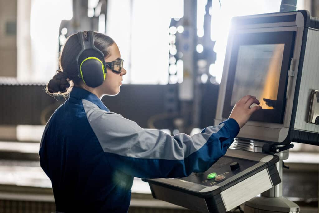 A young woman in safety gear attentively adjusts settings on a CNC water jet cutter, showcasing precision and expertise in an industrial setting.